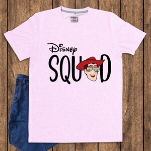 May include: A light pink short-sleeved t-shirt featuring the black text Disney SQUAD with a graphic of a cowboy character wearing a red hat and sunglasses reflecting a castle. The shirt has a crew neckline and a small KandT Apparel brand label.