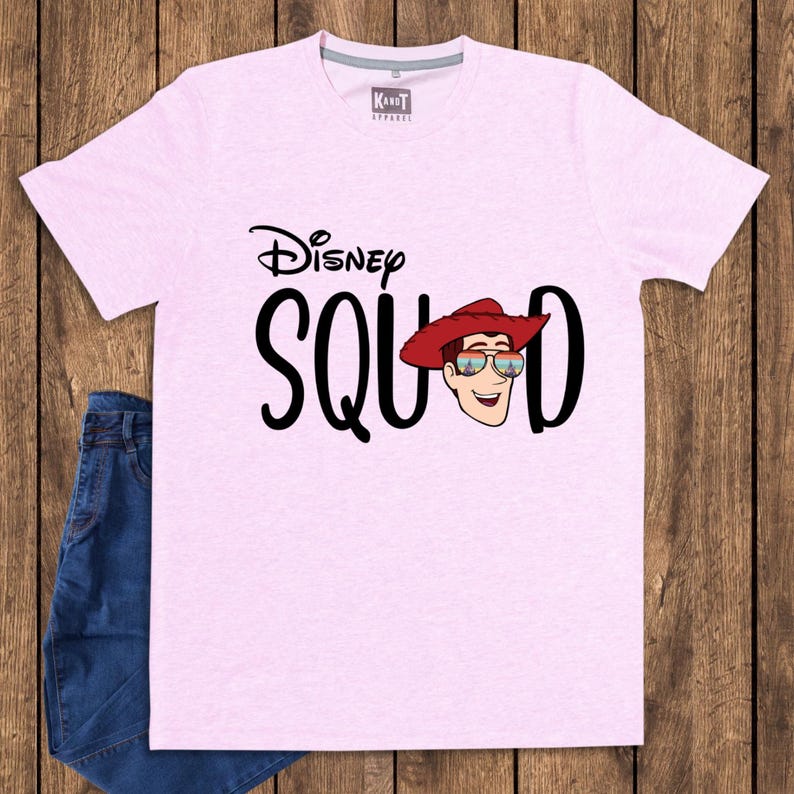 May include: A light pink short-sleeved t-shirt featuring the black text Disney SQUAD with a graphic of a cowboy character wearing a red hat and sunglasses reflecting a castle. The shirt has a crew neckline and a small KandT Apparel brand label.