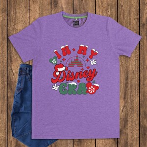 Może przedstawiać: Fioletowa koszulka z napisem "In My Disney Era" w kolorze czerwonym, białym i zielonym. Projekt zawiera zamek, rękawice Myszki Miki i czapkę Świętego Mikołaja. Koszulka jest zestawiona z niebieskimi dżinsami.