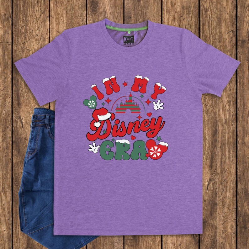 Może przedstawiać: Fioletowa koszulka z napisem "In My Disney Era" w kolorze czerwonym, białym i zielonym. Projekt zawiera zamek, rękawice Myszki Miki i czapkę Świętego Mikołaja. Koszulka jest zestawiona z niebieskimi dżinsami.