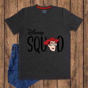 May include: A charcoal gray short-sleeve t-shirt featuring the text Disney SQUAD in black lettering. The letter U is replaced by a cartoon illustration of a character wearing a red cowboy hat and sunglasses reflecting a sunset landscape.