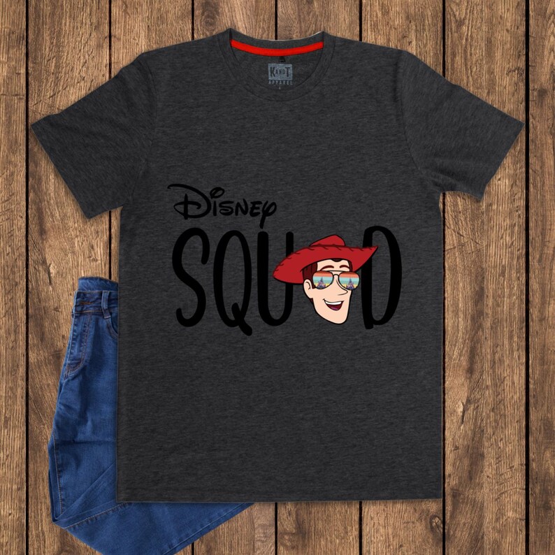 May include: A charcoal gray short-sleeve t-shirt featuring the text Disney SQUAD in black lettering. The letter U is replaced by a cartoon illustration of a character wearing a red cowboy hat and sunglasses reflecting a sunset landscape.