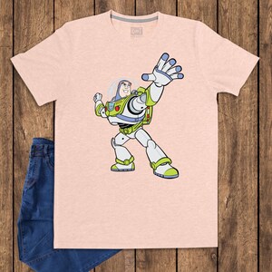 May include: A light pink t-shirt featuring a cartoon illustration of Buzz Lightyear from Toy Story. The character is depicted in his signature spacesuit, with arms outstretched. Blue jeans are visible on the left side of the image.
