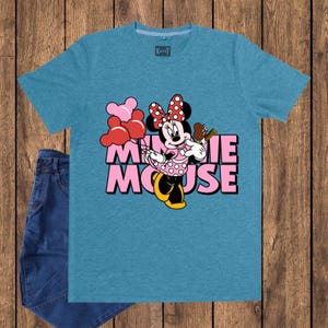 May include: A teal t-shirt featuring a graphic of Minnie Mouse holding balloons and an ice cream bar. The text "MINNIE MOUSE" is printed in pink. A pair of blue jeans is visible on the left side of the image.