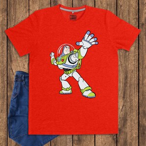 May include: A red t-shirt featuring a cartoon illustration of Buzz Lightyear from Toy Story. The character is depicted in his signature space ranger suit, with a white and green color scheme. The shirt is paired with blue jeans.