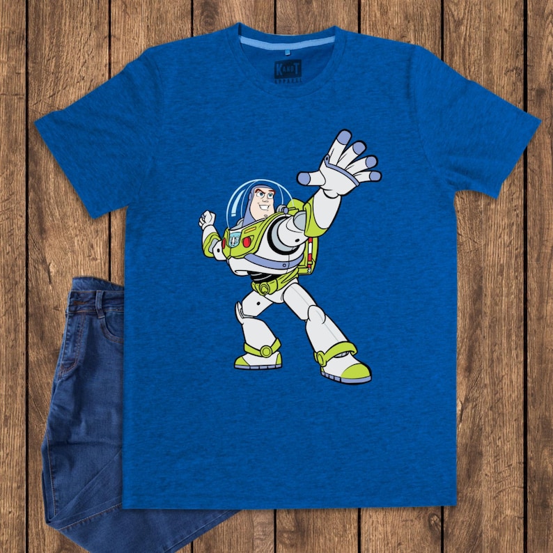 May include: A blue t-shirt featuring a cartoon image of Buzz Lightyear from Toy Story. The character is depicted in his signature white, green, and purple spacesuit, with his arm raised. A pair of blue jeans is next to the shirt.