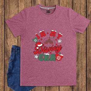 Może przedstawiać: Bordowa koszulka z napisem "IN MY Disney ERA" w świątecznym, kolorowym wzorze. Projekt zawiera czapkę Świętego Mikołaja, rękawice Myszki Miki i sylwetkę zamku. Para niebieskich dżins&oacute;w jest widoczna po lewej stronie.