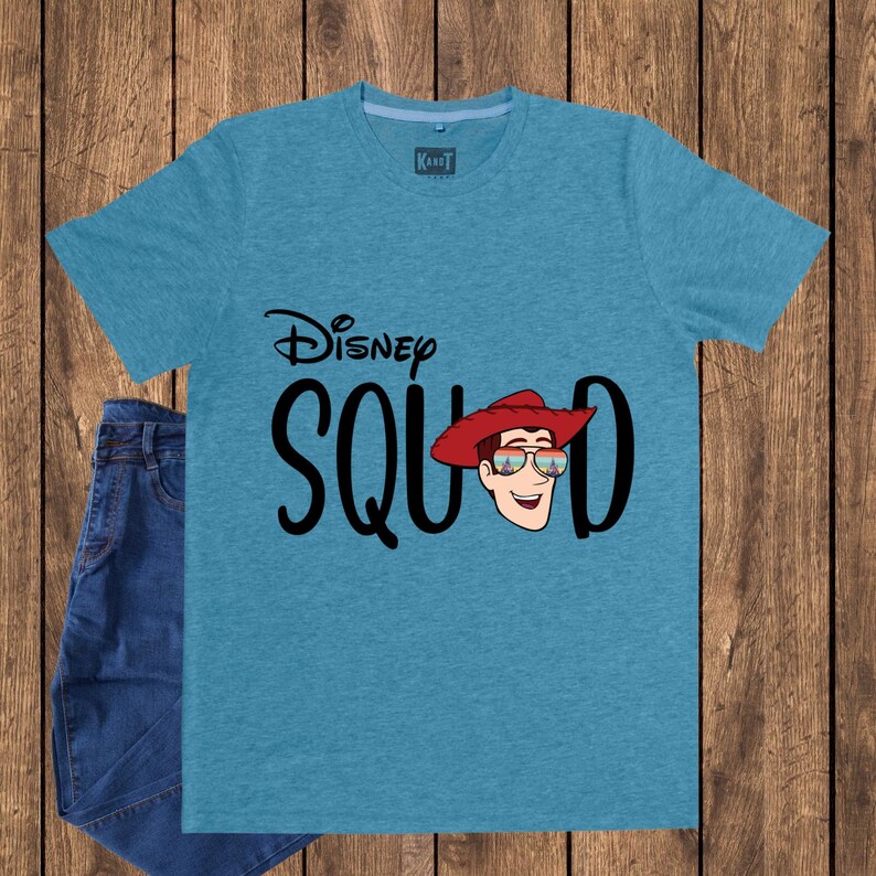 May include: A heather blue short-sleeve t-shirt featuring the black text Disney SQUAD with a graphic of a cowboy character wearing a red hat and sunglasses reflecting a castle. The shirt has a crew neckline and a small brand label at the inner neck.