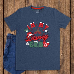Może przedstawiać: Granatowa koszulka z napisem "IN MY Disney ERA" w kolorze czerwonym i zielonym. Projekt zawiera rękawice Myszki Miki, czapkę Świętego Mikołaja i choinkę. Obok koszulki leżą niebieskie dżinsy.