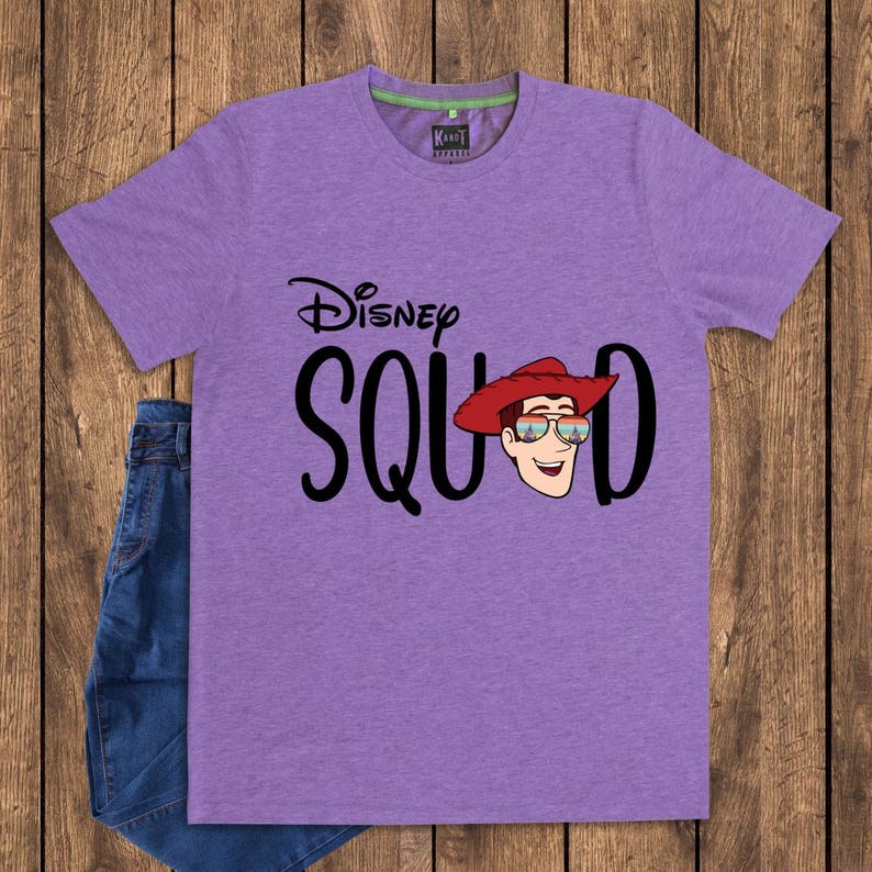 May include: A heather purple short-sleeve t-shirt featuring the black text Disney SQUAD. The letter U is replaced by a graphic of a cowboy character wearing a red hat and sunglasses with a sunset and castle reflection on the lenses.