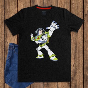 May include: Black t-shirt featuring a cartoon illustration of Buzz Lightyear in his space suit. The character is depicted with a white and green spacesuit, a clear helmet, and is in a dynamic pose with one arm raised. The shirt has a red collar.
