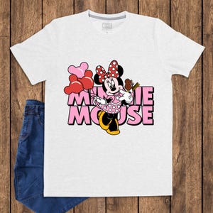 May include: A white t-shirt featuring a graphic of Minnie Mouse holding balloons and a treat. The text "MINNIE MOUSE" is in pink with a black outline. The shirt is paired with blue jeans.