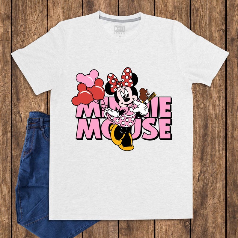 May include: A white t-shirt featuring a graphic of Minnie Mouse holding balloons and a treat. The text "MINNIE MOUSE" is in pink with a black outline. The shirt is paired with blue jeans.