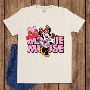 May include: A light beige t-shirt featuring a graphic of Minnie Mouse holding balloons and a chocolate bar. The text "MINNIE MOUSE" is in pink block letters. A pair of blue jeans is visible on the left side of the image.