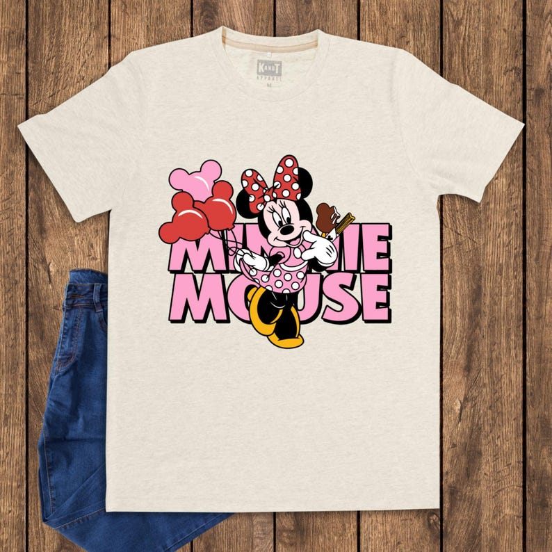 May include: A light beige t-shirt featuring a graphic of Minnie Mouse holding balloons and a chocolate bar. The text "MINNIE MOUSE" is in pink block letters. A pair of blue jeans is visible on the left side of the image.