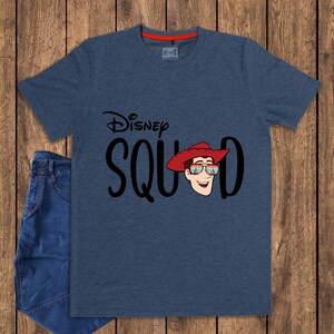 May include: A heathered navy blue short-sleeve t-shirt featuring the black text Disney SQUAD with a graphic of a cartoon character wearing a red cowboy hat and sunglasses. The shirt has a red inner neck trim and is paired with blue denim jeans.