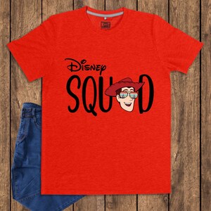May include: A bright red short-sleeved t-shirt featuring the black text Disney SQUAD. The letter U is replaced by a graphic of a cartoon character wearing a red cowboy hat and sunglasses with a sunset reflection. The shirt has a crew neckline.