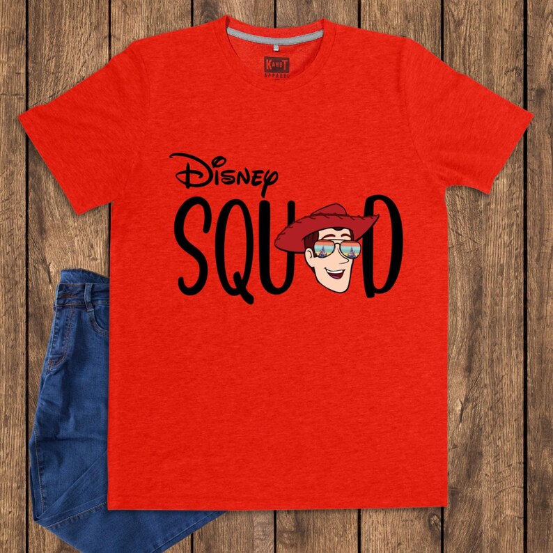 May include: A bright red short-sleeved t-shirt featuring the black text Disney SQUAD. The letter U is replaced by a graphic of a cartoon character wearing a red cowboy hat and sunglasses with a sunset reflection. The shirt has a crew neckline.