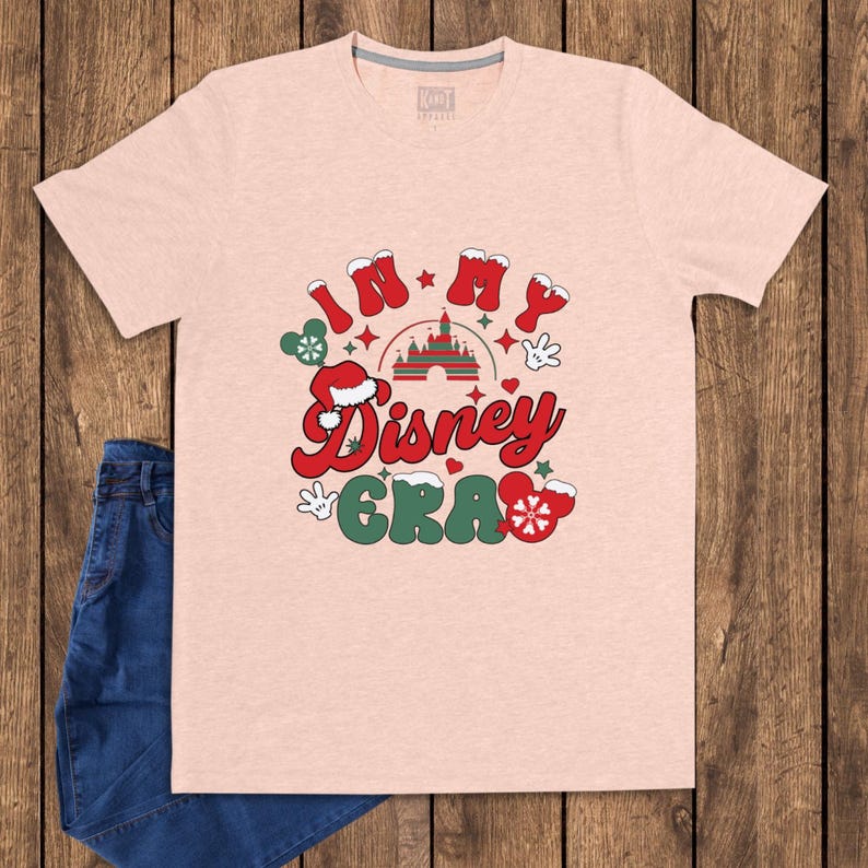 Może przedstawiać: Jasnor&oacute;żowa koszulka z napisem "IN MY Disney ERA" w kolorze czerwonym i zielonym, z grafiką Myszki Miki i zamku. Koszulka jest zestawiona z niebieskimi dżinsami na drewnianym tle.