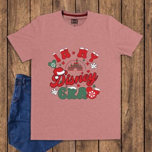 Może przedstawiać: R&oacute;żowa koszulka z napisem "In My Disney Era" w kolorze czerwonym, zielonym i białym. Projekt zawiera rękawice Myszki Miki, zamek i czapkę Świętego Mikołaja. Para niebieskich dżins&oacute;w jest widoczna po lewej stronie.