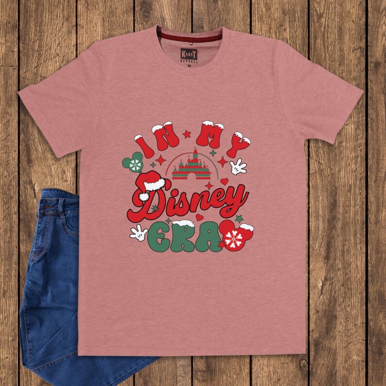 Może przedstawiać: R&oacute;żowa koszulka z napisem "In My Disney Era" w kolorze czerwonym, zielonym i białym. Projekt zawiera rękawice Myszki Miki, zamek i czapkę Świętego Mikołaja. Para niebieskich dżins&oacute;w jest widoczna po lewej stronie.