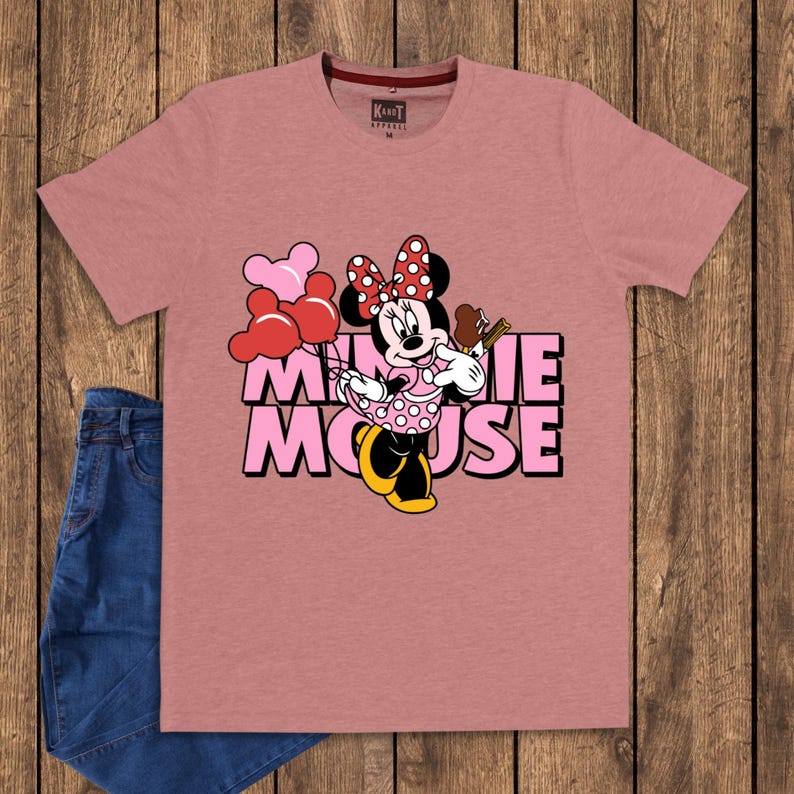 May include: Dusty rose t-shirt featuring a Minnie Mouse graphic. The graphic includes Minnie Mouse holding balloons and a treat, with the words "MINNIE MOUSE" in pink block letters. The shirt is paired with blue jeans.