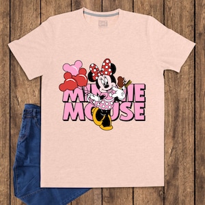 May include: A light pink t-shirt featuring a graphic of Minnie Mouse holding balloons and a chocolate bar. The text "MINNIE MOUSE" is printed in pink with a black outline. A pair of blue jeans is visible on the left side.