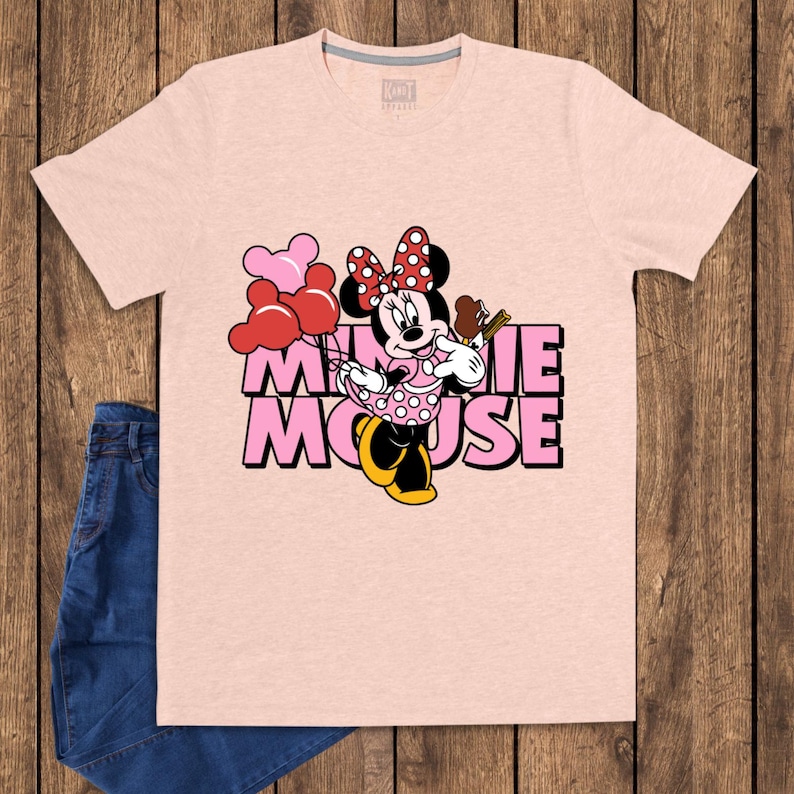 May include: A light pink t-shirt featuring a graphic of Minnie Mouse holding balloons and a chocolate bar. The text "MINNIE MOUSE" is printed in pink with a black outline. A pair of blue jeans is visible on the left side.
