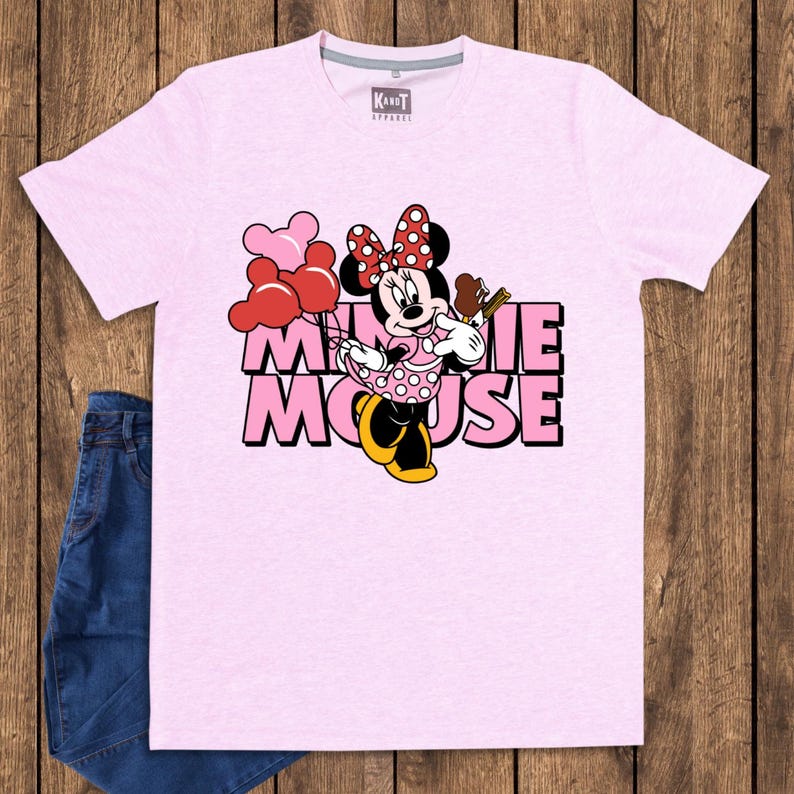 May include: A light pink t-shirt featuring a graphic of Minnie Mouse holding balloons and a treat. The text "Minnie Mouse" is printed in pink and white. A pair of blue jeans is visible on the left side of the image.