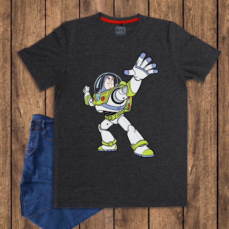 May include: Dark gray t-shirt featuring a cartoon illustration of Buzz Lightyear from Toy Story. The character is depicted in his signature spacesuit, with white, green, and purple accents. Blue jeans are folded next to the shirt.