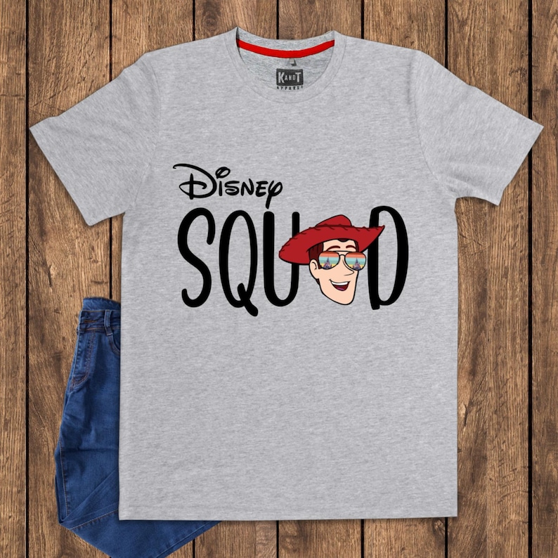 May include: A heather gray short-sleeve t-shirt featuring the black text Disney SQUAD, with the letter U replaced by a cartoon illustration of a character wearing a red cowboy hat and sunglasses reflecting a castle and sunset landscape.