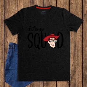 May include: A charcoal gray short-sleeve t-shirt featuring the black text Disney SQUAD with a graphic of a cartoon character wearing a red cowboy hat and sunglasses reflecting a castle scene. The shirt has a red inner collar detail.