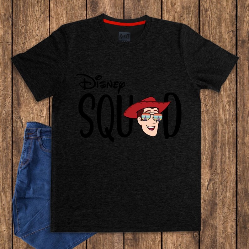 May include: A charcoal gray short-sleeve t-shirt featuring the black text Disney SQUAD with a graphic of a cartoon character wearing a red cowboy hat and sunglasses reflecting a castle scene. The shirt has a red inner collar detail.