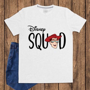 May include: A light gray short-sleeve t-shirt featuring the black text Disney SQUAD with the letter U replaced by a cartoon illustration of a character wearing a red cowboy hat and sunglasses reflecting a castle and fireworks display.