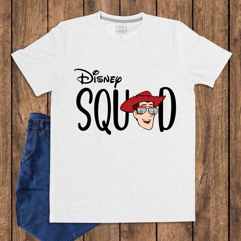 May include: A light gray short-sleeve t-shirt featuring the black text Disney SQUAD with the letter U replaced by a cartoon illustration of a character wearing a red cowboy hat and sunglasses reflecting a castle and fireworks display.
