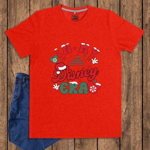 Może przedstawiać: Czerwona koszulka z napisem "IN MY Disney ERA" w stylu retro. Projekt zawiera czapkę Świętego Mikołaja, płatki śniegu i dłonie Myszki Miki. Para niebieskich dżins&oacute;w jest widoczna po lewej stronie obrazu.