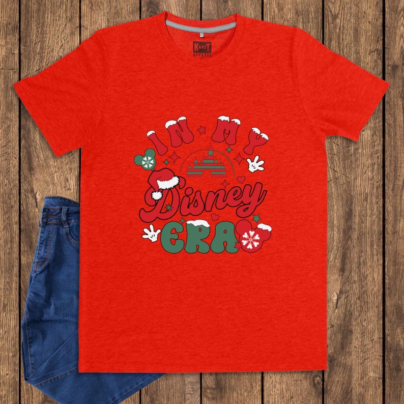 Może przedstawiać: Czerwona koszulka z napisem "IN MY Disney ERA" w stylu retro. Projekt zawiera czapkę Świętego Mikołaja, płatki śniegu i dłonie Myszki Miki. Para niebieskich dżins&oacute;w jest widoczna po lewej stronie obrazu.