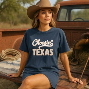 May include: A navy blue t-shirt with the text "Choosin' TEXAS" in a cream-colored, vintage-style font. The person in the image is wearing a brown cowboy hat. The background includes a rusty red truck bed and a coiled rope.