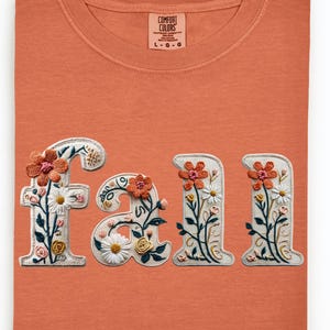 May include: An orange Comfort Colors t-shirt with the word "fall" embroidered in white with floral accents. The flowers are in shades of orange, pink, and white, with green leaves. The shirt is folded neatly.