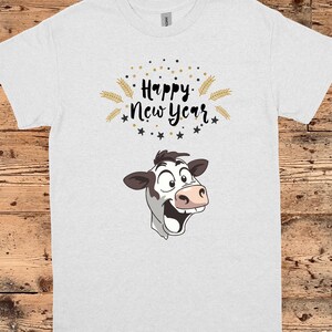 Puede incluir: Camiseta blanca con una vaca de dibujos animados sonriendo. La camiseta tiene el texto "Happy New Year" en escritura negra con detalles dorados, estrellas y espigas de trigo.