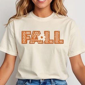 May include: Cream-colored t-shirt with the word "FALL" in large, decorative, orange letters. The letters have a detailed, patterned design, with floral and swirl motifs. The shirt is a casual, short-sleeved style.