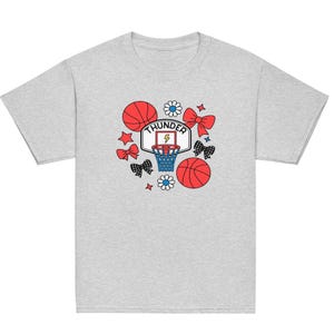 Op de afbeelding: Lichtgrijs t-shirt met een basketbalthema. Het ontwerp bevat rode basketballen, strikken en sterren, een basketbalring en het woord "THUNDER". Het ontwerp is in rood, wit en blauw.