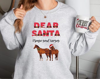 May include: A light gray sweatshirt with the text "DEAR SANTA Please send horses" in red and black. The design includes illustrations of Santa's belt and two brown horses, one wearing a Santa hat. A white mug with "Merry Christmas" is held in the hand.