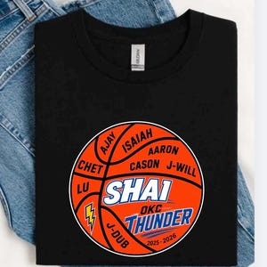 Op de afbeelding: Zwart t-shirt met een basketbalontwerp. Het ontwerp bevat namen van spelers en de tekst "SHAI OKC THUNDER 2025-2026" in oranje, blauw en wit. Het t-shirt wordt getoond op een denim achtergrond.