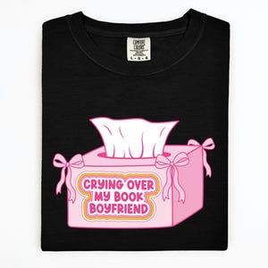 Puede incluir: Camiseta negra con un diseño de caja de pañuelos rosa y el texto "Crying Over My Book Boyfriend". La caja tiene lazos. La camiseta doblada tiene una etiqueta Comfort Colors.