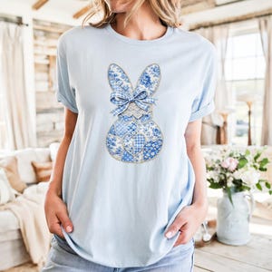 May include: Light blue t-shirt featuring a patchwork bunny design. The bunny is composed of various blue and white floral and gingham patterns, with a lace-trimmed bow. The shirt is a short-sleeved crew neck.