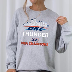May include: A light gray sweatshirt featuring the text "OKC THUNDER 2025 NBA CHAMPIONS" in white, blue, and orange. Above the text is a skyline graphic with player names. The long-sleeved top is made of a soft material.