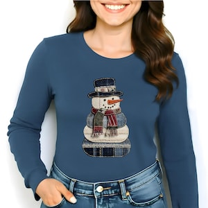 May include: A long-sleeved, blue shirt featuring a snowman applique. The snowman has a black top hat, a carrot nose, and a scarf with red and green stripes. The snowman is made of denim and cream-colored fabric.