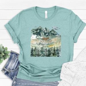 May include: A light teal t-shirt featuring a watercolor design of mountains and trees. The shirt has the text "How beautiful on the mountains are the feet of those who bring good news... Isaiah 52:7."