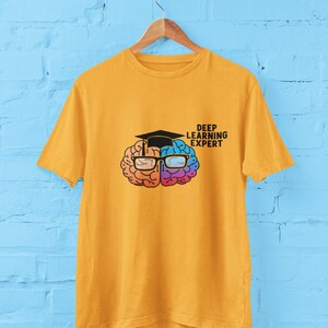 Puede incluir: Una camiseta amarilla con un gráfico de un cerebro que lleva una gorra de graduación y gafas. El texto "Deep Learning Expert" está impreso en la camiseta.